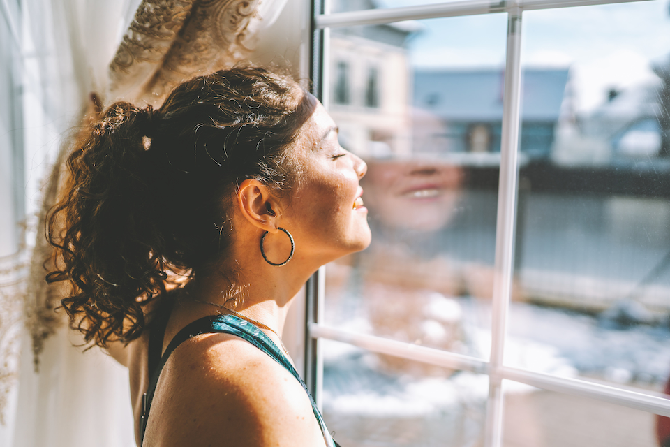 Middle age woman wearing sportswear is standing by a large window and exposing her face to the sun.