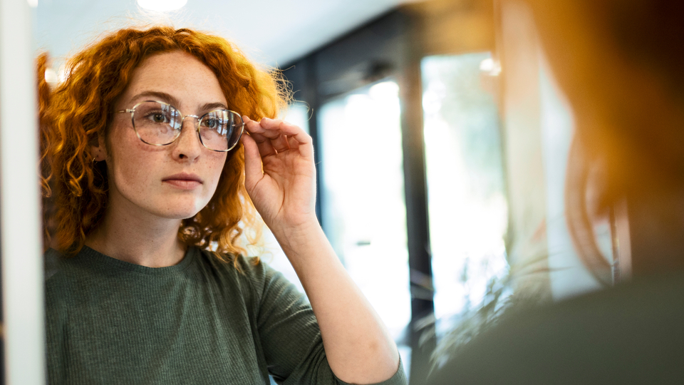 A young woman with red curly hair trying on glasses at the optometrist and wearing a jade green top.