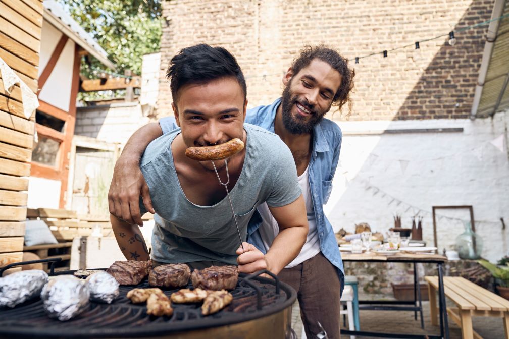 Two people preparing meat for a barbecue in a backyard patio. One person is holding a sausage on a fork and positioning it like a smile.