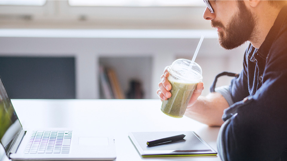 A bearded man sipping a green juice and looking at his laptop