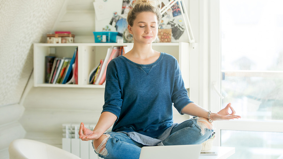 A young woman meditates at home in front of a laptop