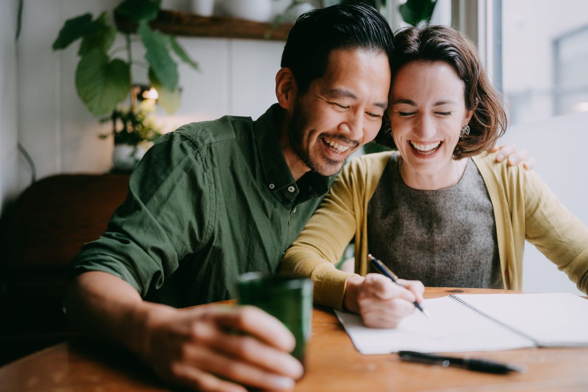 Happy couple sitting next to each other at a table writing in a notebook,