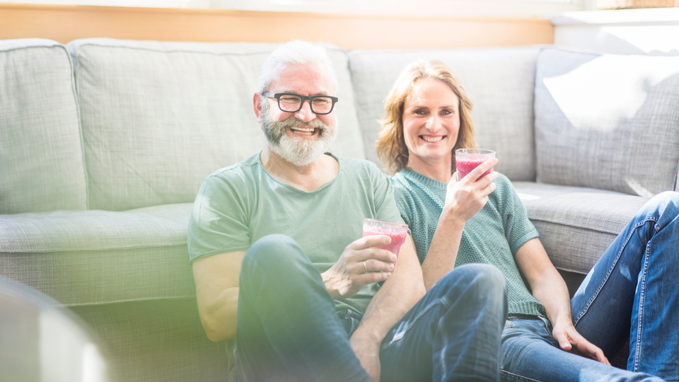 Senior couple sitting on the floor of a loungeroom smiling and drinking juice.