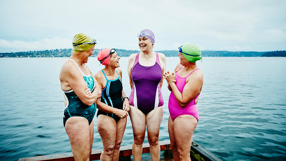 A group of friends in their swimmers, caps and goggles laugh as they stand together