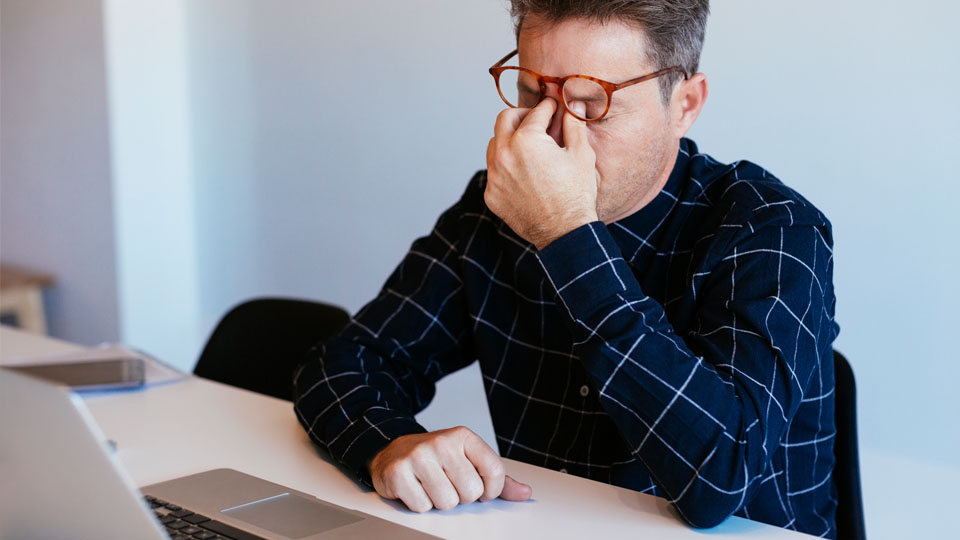 Man pinching his nose, appearing stressed, sitting in front of a laptop.