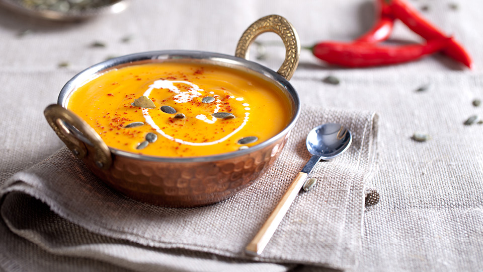 Pumpkin soup in a metal bowl on a table setting with chilis in the background.