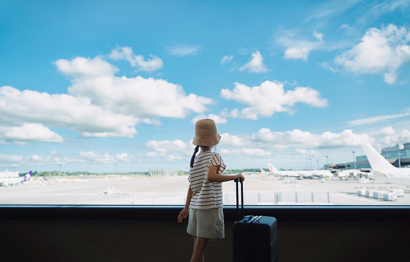 A young girl looks out a large window at an airport while standing beside her luggage