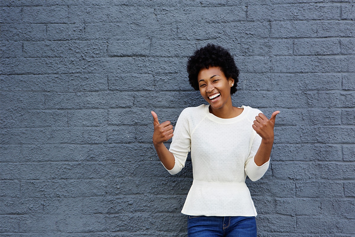 Person standing against a grey brick wall, with two thumbs up, smiling.