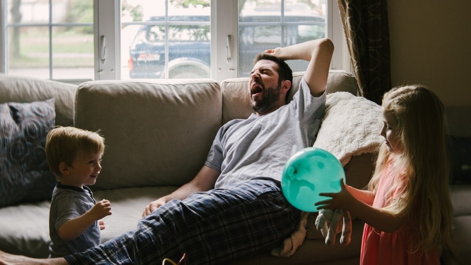 A dad sitting on the couch yawning, while his young son and daughter play with a balloon