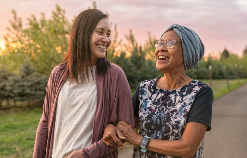 Two smiling women walking arm-in-arm outdoors at sunset, one wearing a pink cardigan, the other in a floral top and blue headwrap.
