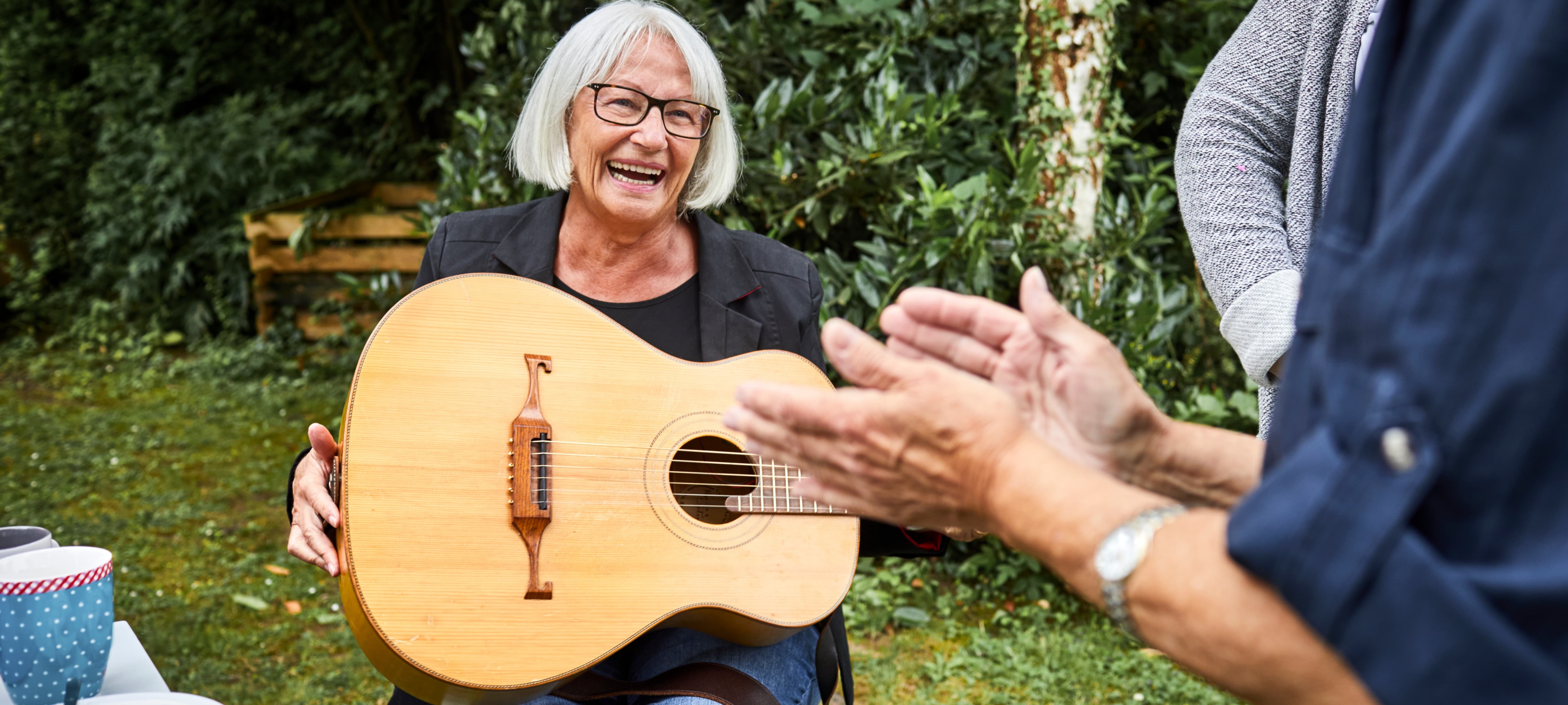 A grey-haired woman in her 70s laughing with a friend while holding an acoustic guitar in her yard.