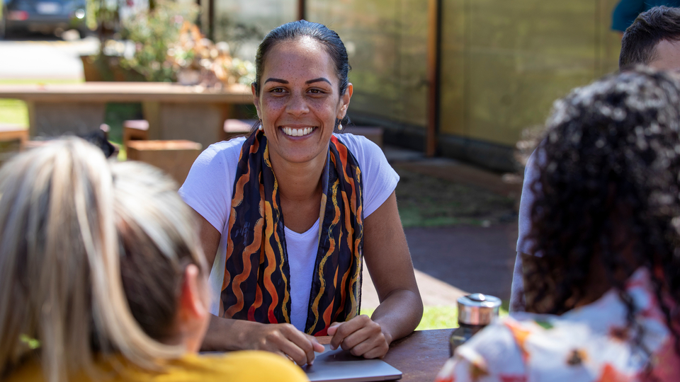 Woman with scarf around her neck smiling at outdoor table, talking to people.