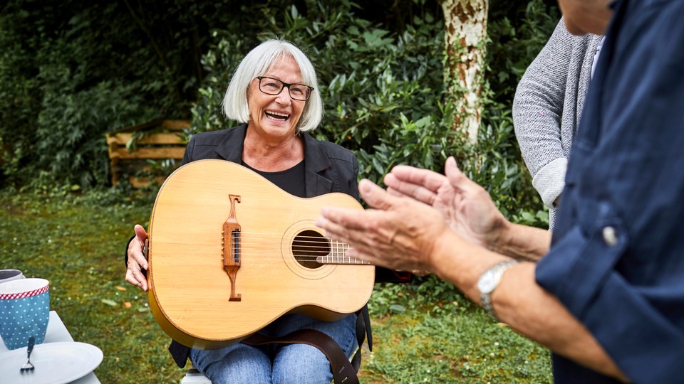 Senior woman playing a guitar, smiling, while people clap in the foreground.