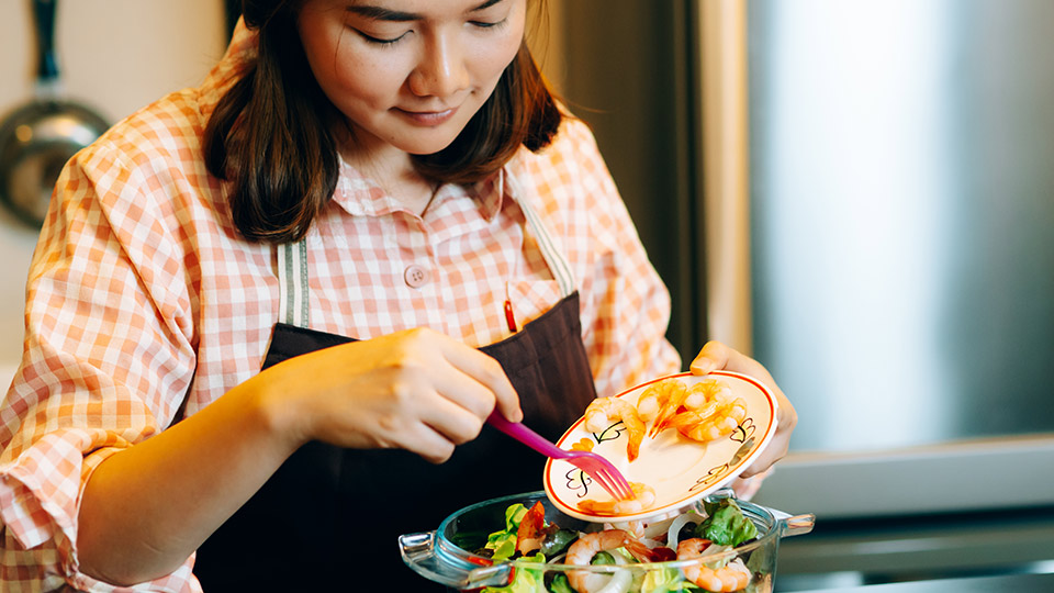 A young woman wearing an apron adds prawns to a colourful salad