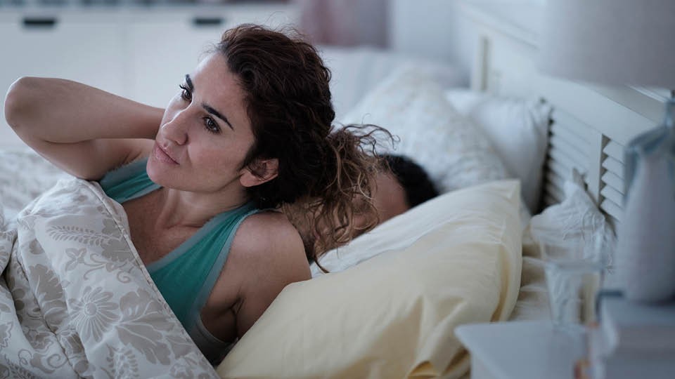 A woman in bed holding her neck with her partner asleep next to her.