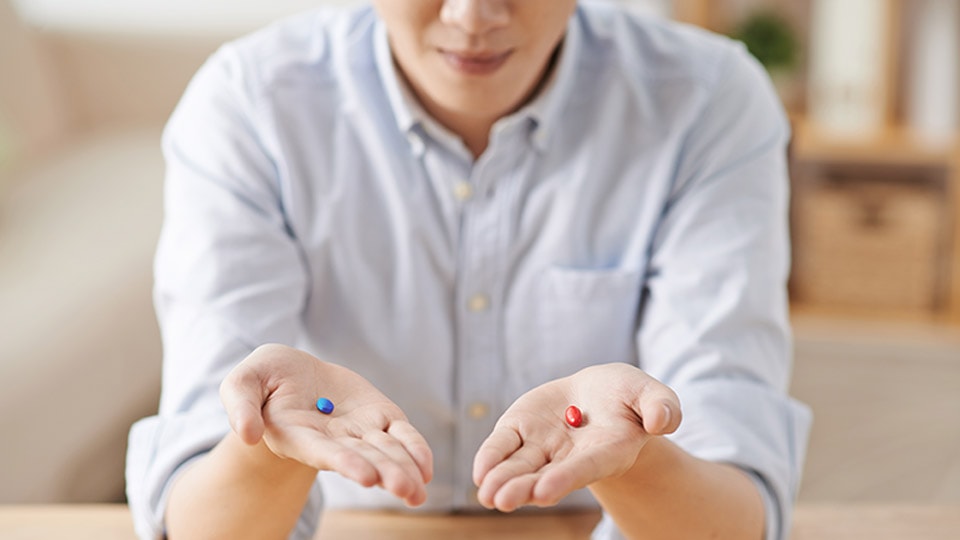 A person holding a red pill in one had and a blue pill in their other hand.