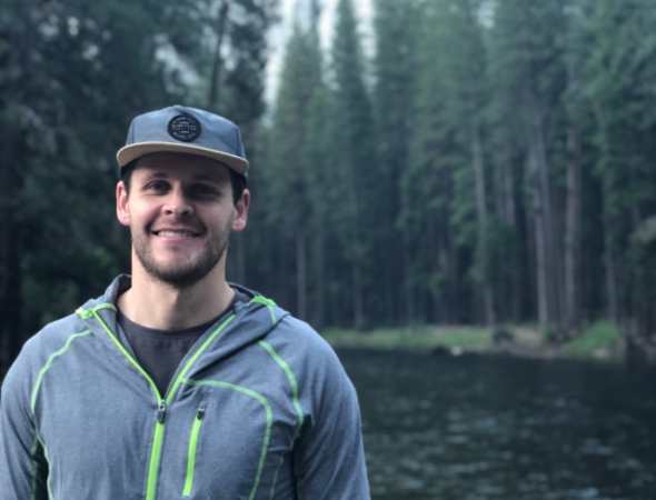 Close up photo of a man wearing a cap with a forest behind him.