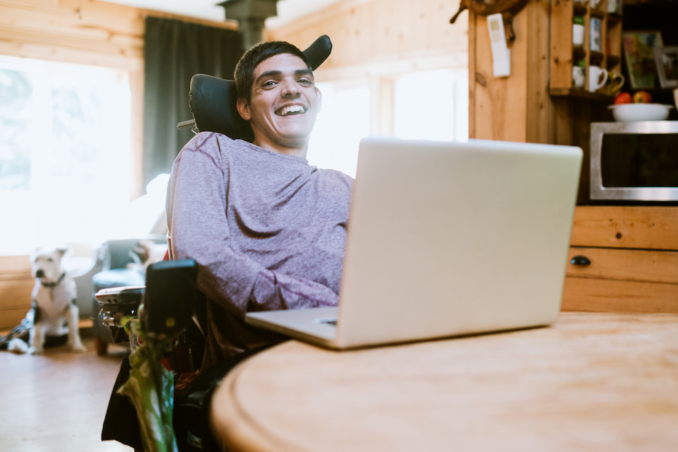 A person with a disability who is in a wheelchair, looking at the camera and laughing, at a table with a laptop.