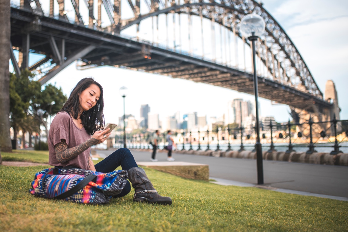 A woman sitting in a park on her phone under the Sydney Harbour Bridge.