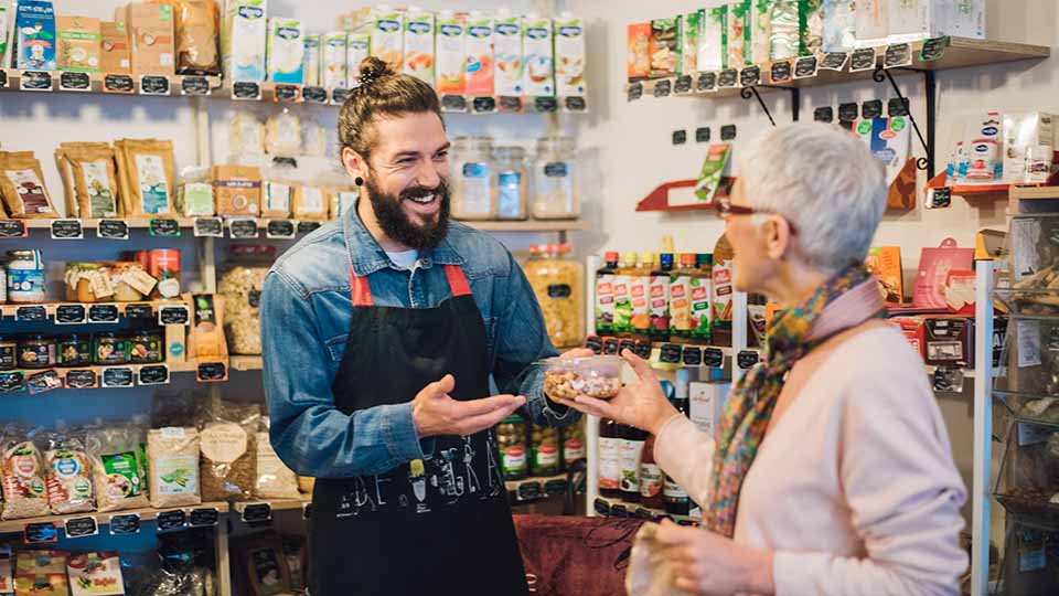 Man wearing an apron in a food store handing an item to a woman shopping.