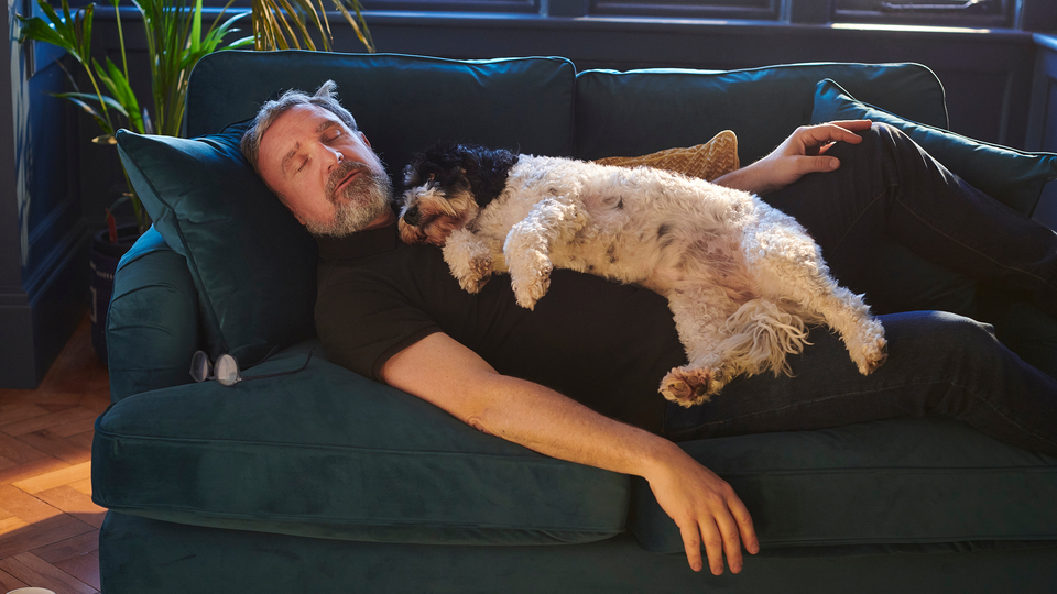 Man with grey hair and beard napping on a jade-coloured couch with his puppy.