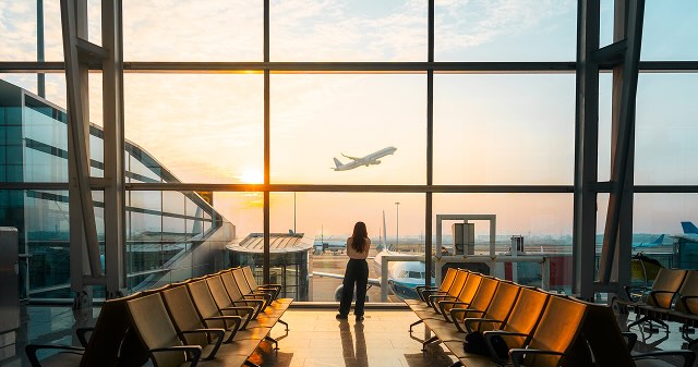A woman standing at an airport window watching a plane take off at sunrise