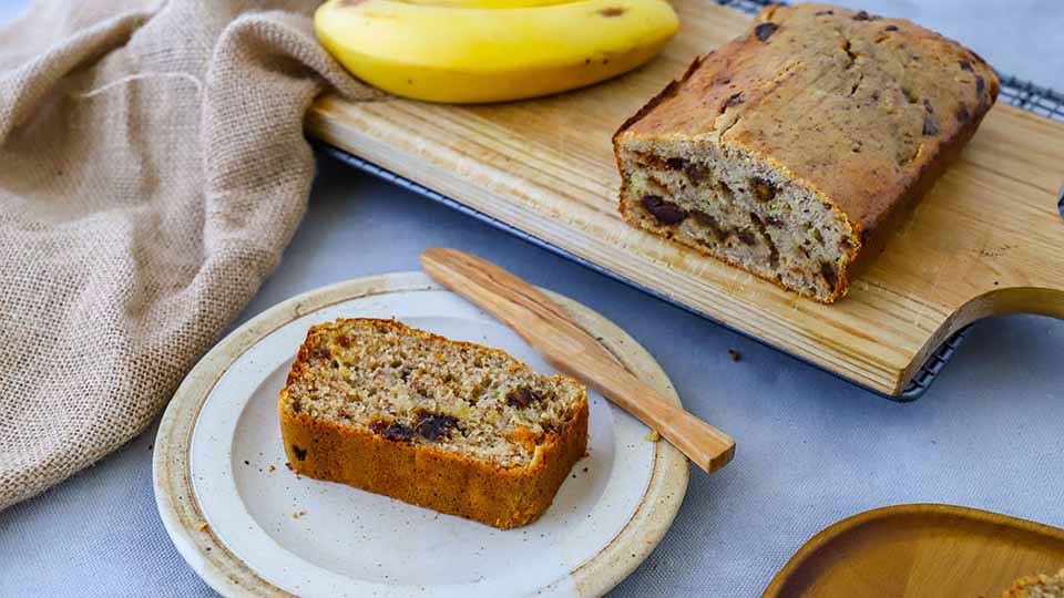 Banana bread on a wooden serving paddle, with a piece sliced off and being served on a saucer.