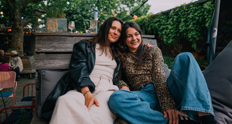Two smiling women sitting close to each other on an outside chair.