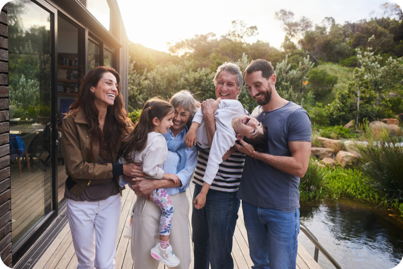 A multi-generation family laughing and hugging on a deck outside.