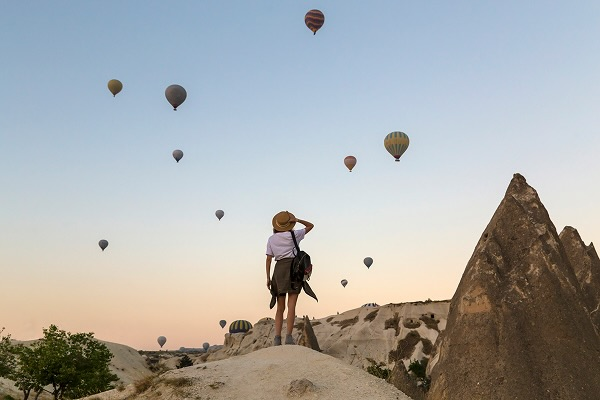 A woman standing on a hill with hot air balloons visible in the air in front of her