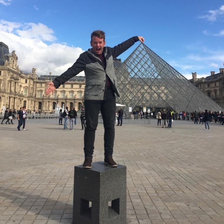 A picture of a man standing in front of the famous Louvre pyramid