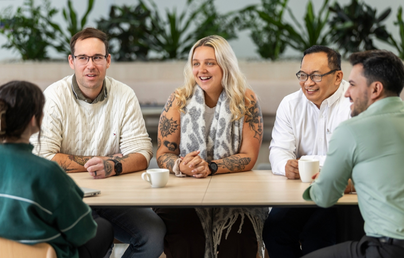 Five diverse professionals smiling during a meeting around a table in a modern office with plants in the background.