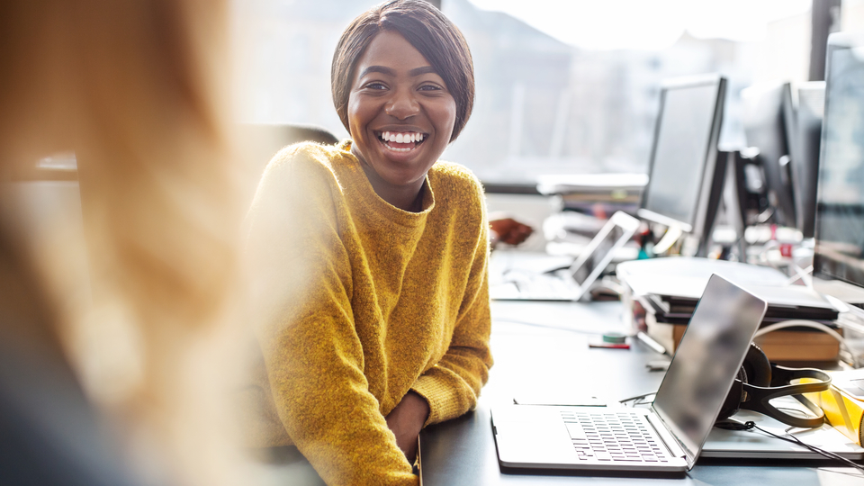 Person sitting at a desk with a laptop, turned towards another person, smiling.