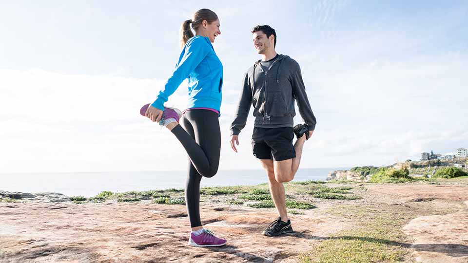 Two people wearing activewear outdoors, doing leg stretches.