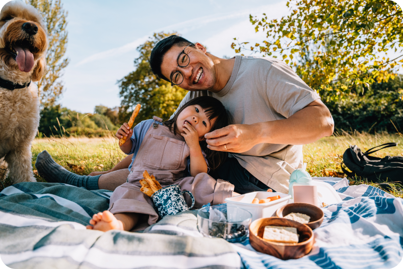 A man, a toddler and a dog having a picnic together outside.