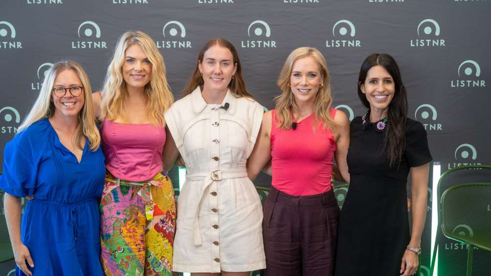 Five women stand arm-in-arm posing for a group photo in front of a media wall.