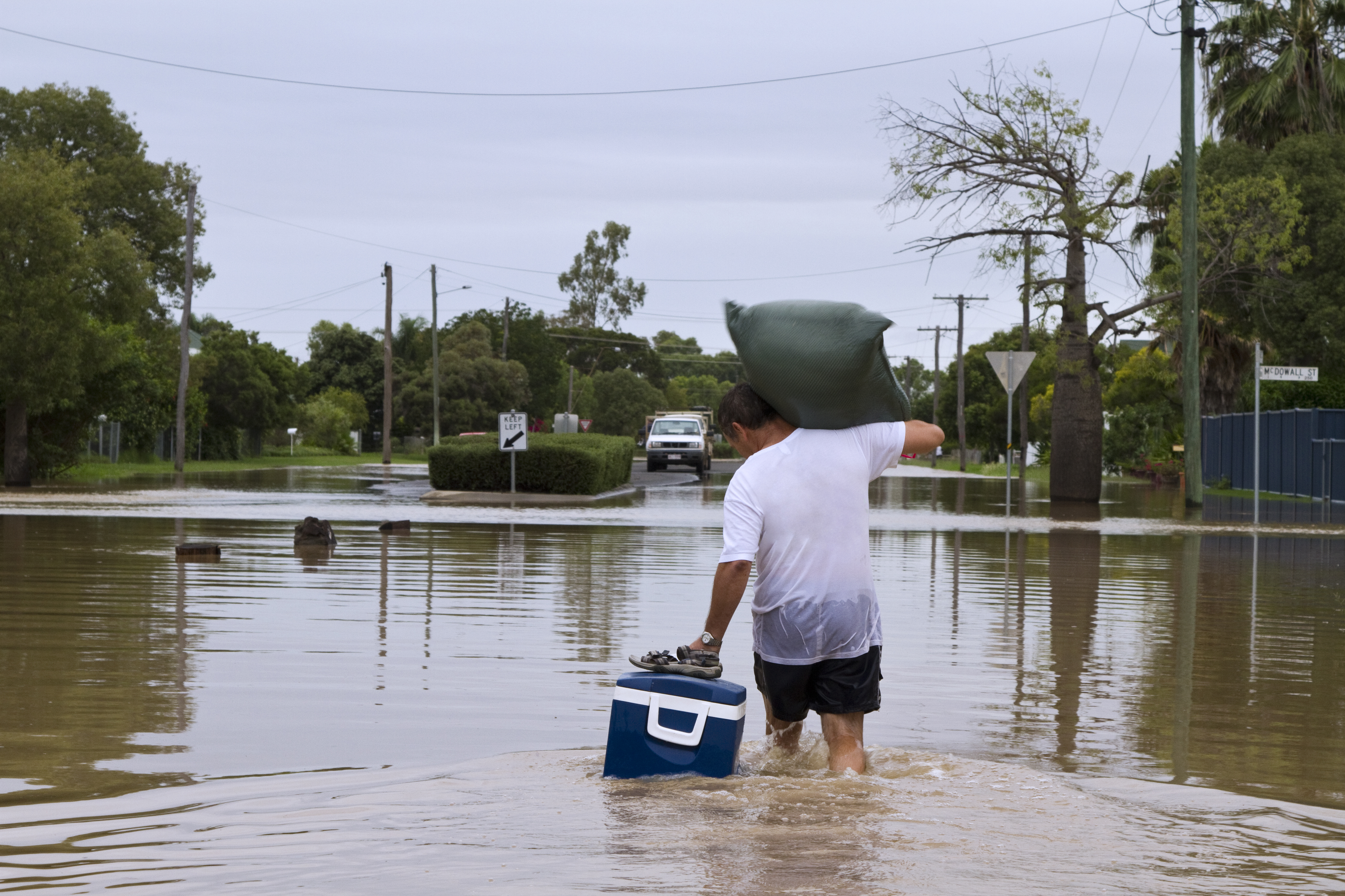 Man dragging an eski through a flooded street and a garbage bag on his right shoulder