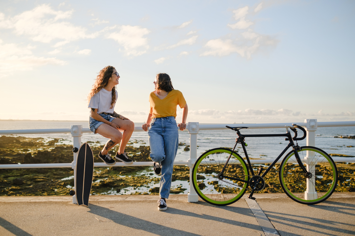 Two friends with a bike and skateboard talking in front of the sea.