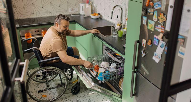 A man in a wheelchair opening a dishwasher in a kitchen.