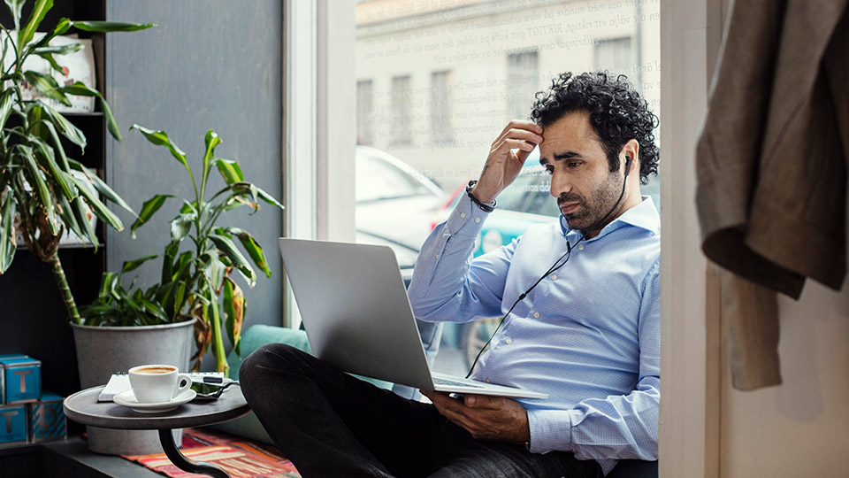 A man wearing business attire scratches his forehead as he sits in front of his laptop with headphones plugged in