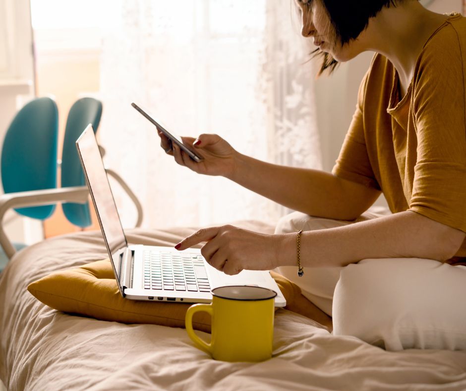 A woman sitting on her bed using her laptop and phone.