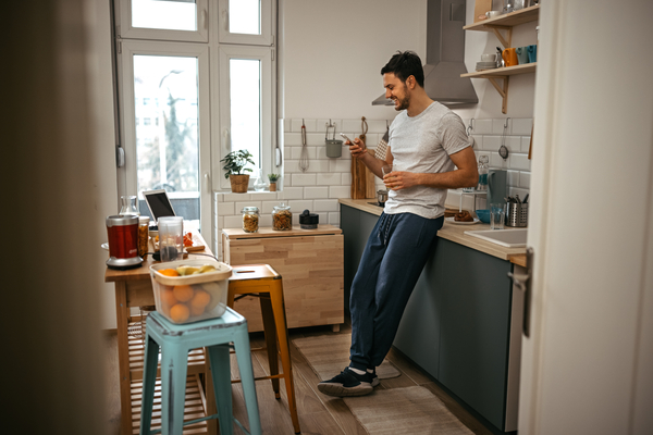 A man standing in his kitchen, looking at his phone while smiling