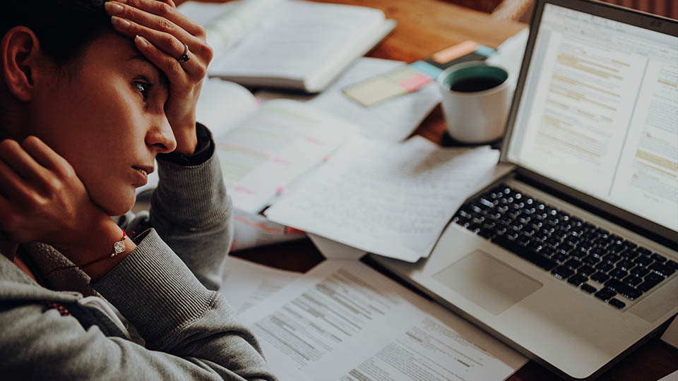 A stressed woman holds her head in her hands as she stares at her laptop screen while surrounded by papers