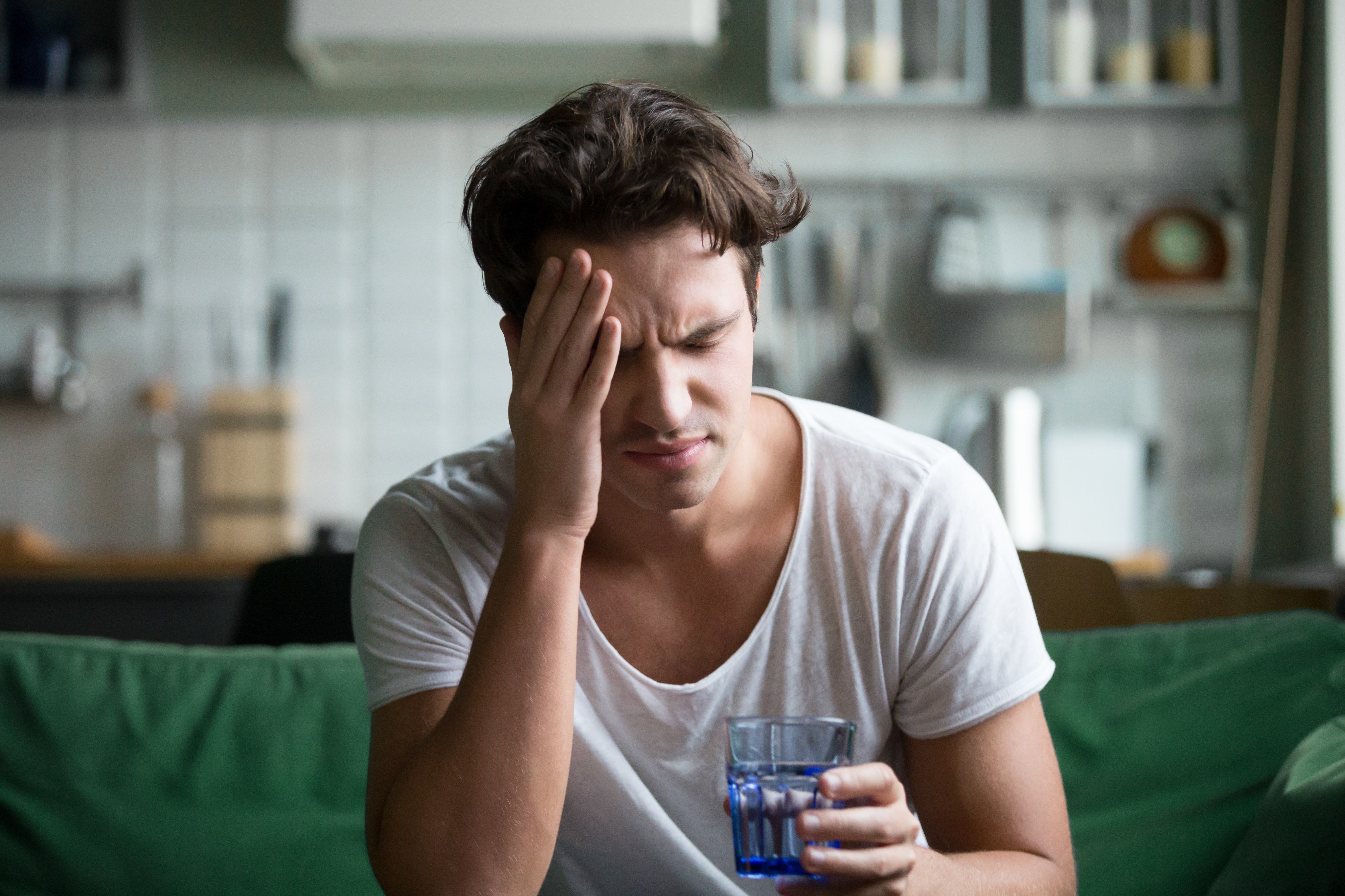 Young man in white t-shirt holding glass of water, touching forehead with pained expression, sitting on green couch in kitchen.