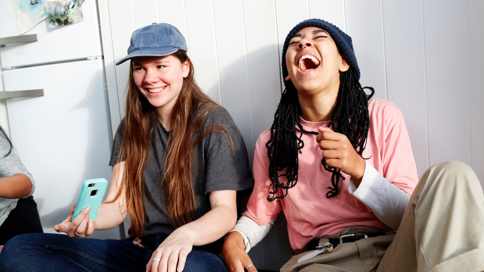 Two young woman sitting against a wall laughing candidly.