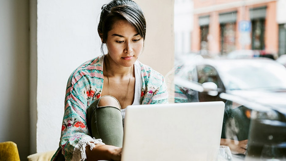 Young person sitting in a window seat using a laptop with a street view behind them.