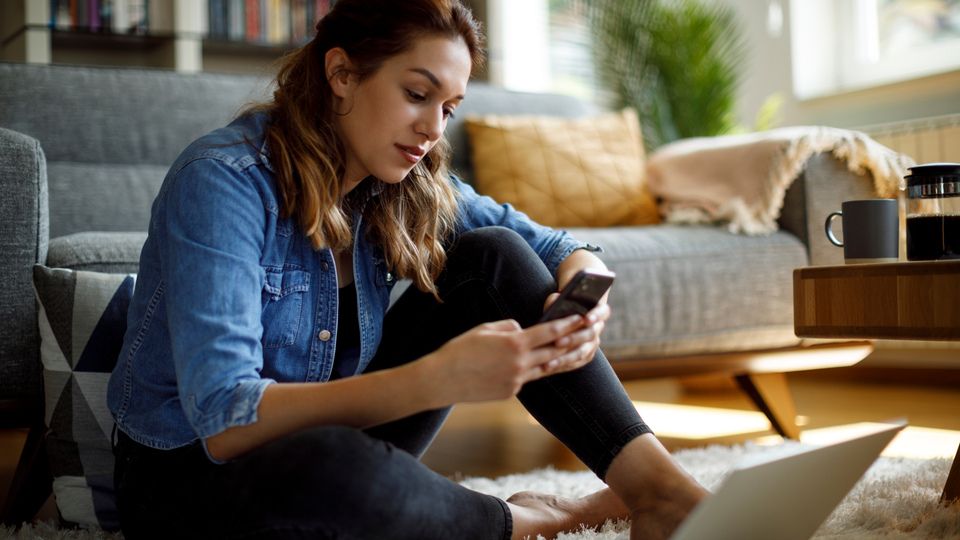 Woman sitting on floor scrolling through phone with open laptop in front of her.