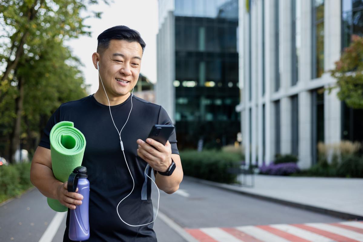 A man walking through a business district holding a yoga mat, water bottle and looking at his smartphone.