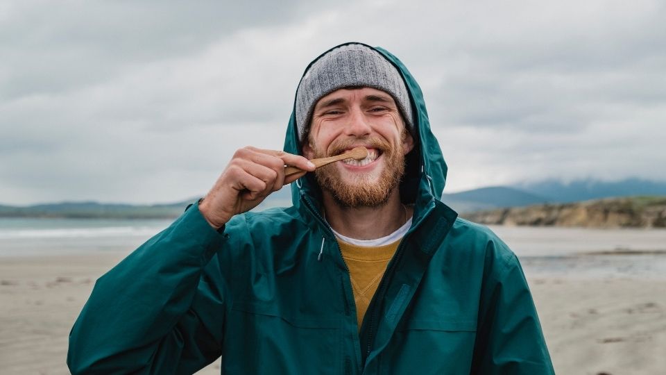 A man wearing a beanie and weatherproof jacket smiles as he brushes his teeth.