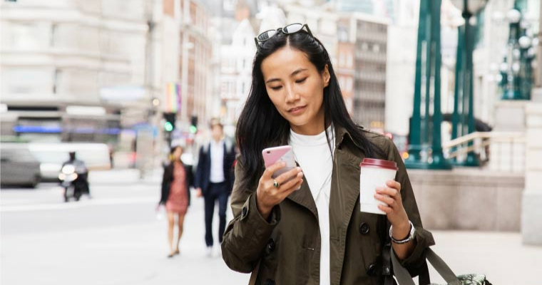A woman walking while looking at her phone and holding a coffee